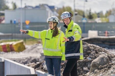 En kvinna och en man i reflexjackor och hjälpar pekar åt varsitt håll på en byggarbetsplats.