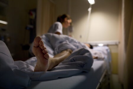En sjuksköterska pysslar om en patient liggande i sin säng med nedtonad belysning. I bildens förgrund syns patientens fot.