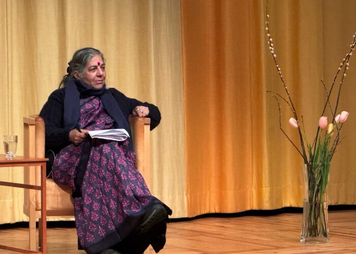 Vandana Shiva klädd i lila sitter på en scen framför ett gult draperi, med en bukett blommor till höger i bild.
