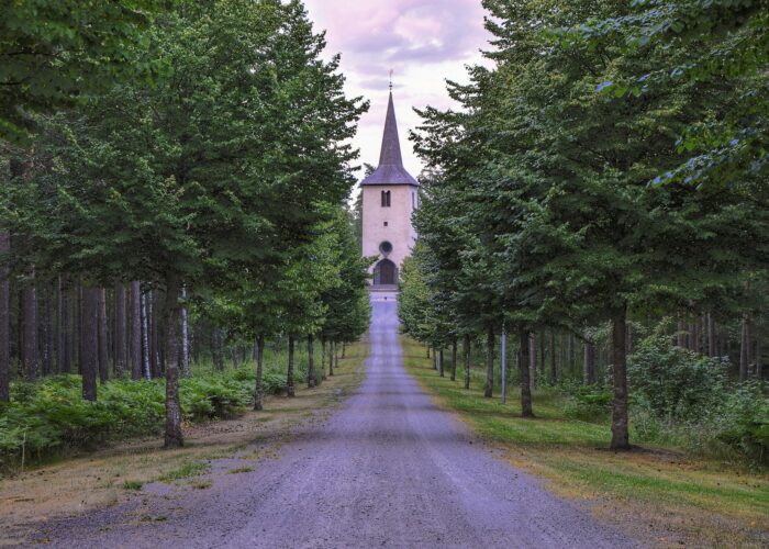 Vit kyrka med spetsigt tak syns i mitten av bilden, en rak väg kantad av skog leder dit.