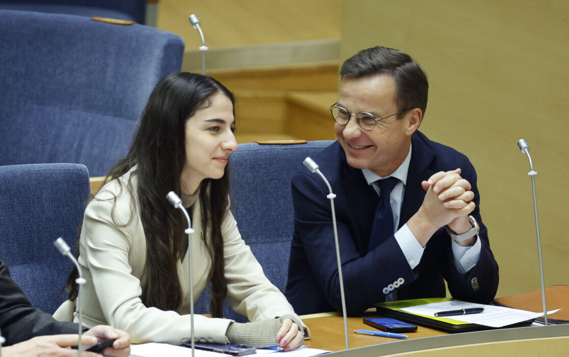 Romina Pourmokhtari och Ulf Kristersson på varsin stol bredvid varandra i riksdagen, leende.
