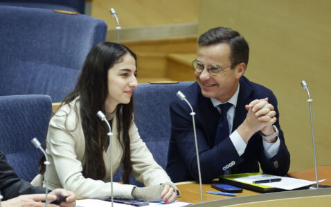 Romina Pourmokhtari och Ulf Kristersson på varsin stol bredvid varandra i riksdagen, leende.