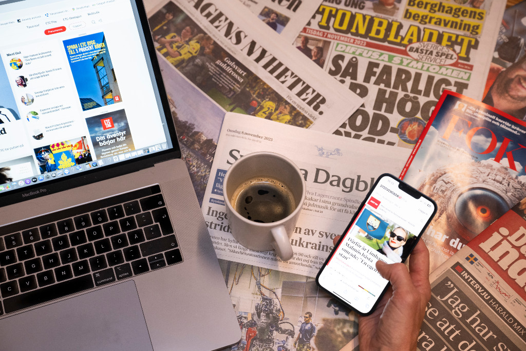 Kaffekopp står på flera dagstidningar invid en datorskärm och mobilskärm.